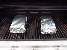 two aluminum foil wrapped hot dogs cooking on an outdoor bbq grill with three knobs