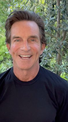 a smiling man wearing a black t - shirt in front of some trees and bushes