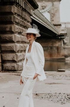 a woman in a white suit and hat standing on the sidewalk with her hands behind her back