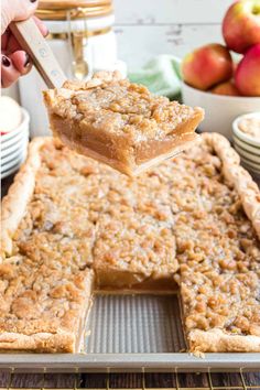 a person holding a piece of apple pie over a baking pan with apples in the background