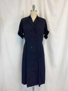 "Vintage 1950s dress featuring; ~ a v neck with notched collar short sleeves with winged cuff lightly padded shoulders collar and cuff are trimmed in velvet has two rows of buttons all the way down the front string belt loops at the waist but no belt skirt is fairly straight and falls to below the knee ~ of a mid weight faille fabric in a deep navy blue ~ labeled \"Make mine a McKettrick\" this has no size label, measurements when laid out flat are: 23 inches from pit to pit 10 inch sleeve 2 1/2 1930s Fashion Women Dresses, Classic Collared Dress With Cuffed Sleeves, Classic Formal Dresses With Cuffed Sleeves, Vintage V-neck Dress For Workwear, Retro Navy Dress For Formal Occasions, Navy Retro Dress For Formal Occasions, Navy Retro Formal Dress, 1950s Style Short Sleeve Vintage Dress For Formal Occasions, Classic Short Sleeve Lined Vintage Dress