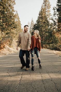 an engaged couple holding hands and walking down the road in front of some pine trees