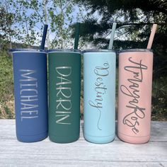 three different colored tumblers with straws sitting on a wooden table outside in front of some trees