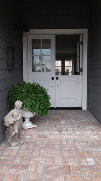 a teddy bear sitting in front of a white door on a brick walkway next to a potted plant