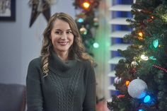 a woman standing in front of a christmas tree with lights on it and smiling at the camera