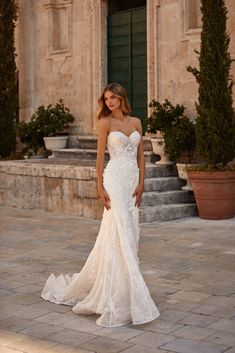 a woman in a white wedding dress standing on a stone walkway with potted plants behind her