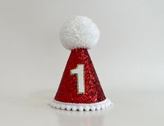 a red and white party hat with the number one on it's side, decorated with pom - poms