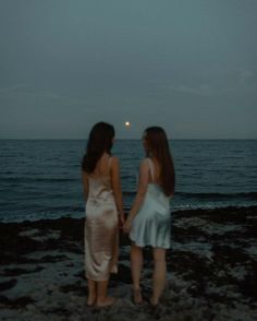 two women standing on the beach holding hands and looking out at the ocean under a full moon