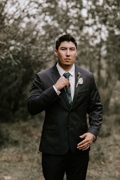 a man wearing a suit and tie standing in front of trees with his hands on his chest