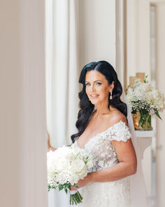 a woman in a wedding dress holding a bouquet and smiling at the camera while standing next to a window