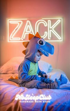 a small child in a dinosaur costume sitting on a bed with a neon sign behind him