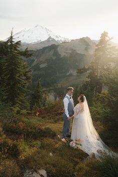 a bride and groom standing on top of a mountain looking at each other while the sun is setting