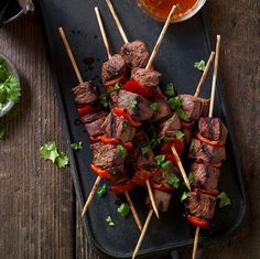 several skewers of meat and vegetables on a black tray with dipping sauces