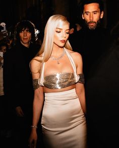 a woman with blonde hair wearing a white dress and gold choker standing next to a man