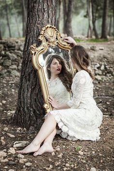 a woman sitting on the ground in front of a tree looking at her reflection in a mirror