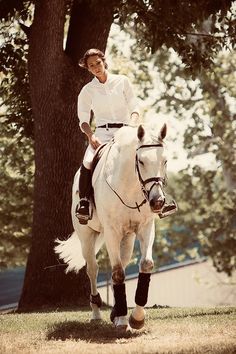 a woman riding on the back of a white horse in front of a large tree