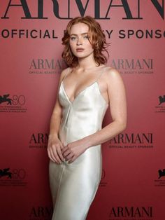 a woman standing in front of a red wall wearing a white dress and posing for the camera