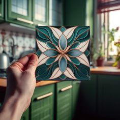 a hand holding up a decorative tile in a green and white kitchen with wooden cabinets