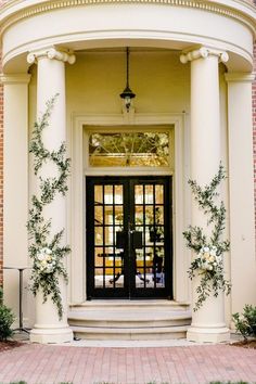 an entrance to a building with two black doors and flowers on the front door, surrounded by greenery