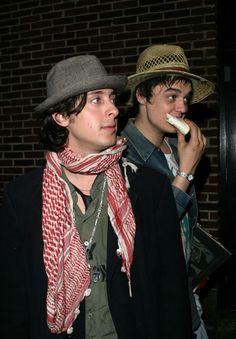 two young men standing next to each other with hats and scarves around their necks