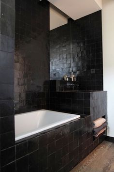 a black tiled bathroom with a white bathtub and wooden flooring, along with dark tile walls