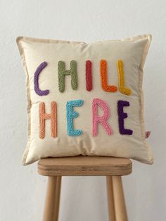 a pillow with the words chill here on it sitting on a wooden stool next to a white wall