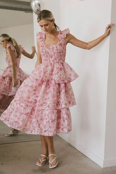 The Absedee Floral Midi Dress is crafted with a smocked bodice that complements its scoop neckline. It boasts charming ruffled cap sleeves and a gracefully flowing three-tiered skirt. The dress is designed with a printed tulle shell that adds a delicate touch, and it is fully lined to ensure both comfort and coverage. Smocked Tiered Skirt Dresses For Garden Party, Knee-length Smocked Dress With Ruffles For Garden Party, Pink Tiered Smocked Dress With Floral Print, Pink Ruffled Smocked Midi Dress, Pink Smocked Midi Dress With Ruffles, Pink Midi Smocked Dress With Ruffles, Spring Smock Dress With Ruffled Straps, Pink Tiered Skirt Dress For Daywear, Spring Dresses With Smock Detail And Ruffled Straps