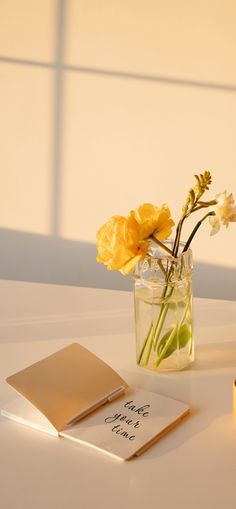 yellow flowers in a glass vase on a white table with notepad and candle next to it