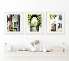 three framed pictures hang on the wall above a white shelf with books and vases