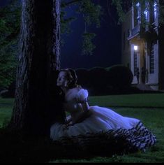 a woman sitting under a tree in front of a house at night with the light on