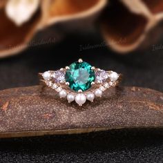 a green ring with pearls and stones on top of a piece of wood next to flowers