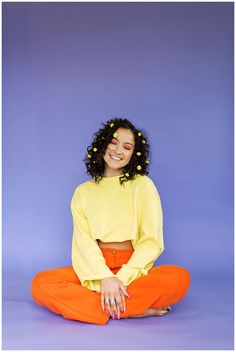 a woman sitting on the ground wearing an orange pants and a yellow top with flowers in her hair