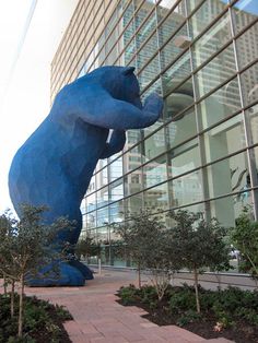 a large blue bear statue sitting in front of a tall building next to trees and bushes