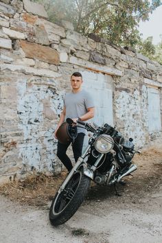 a man standing next to a motorcycle in front of a stone wall with trees on it