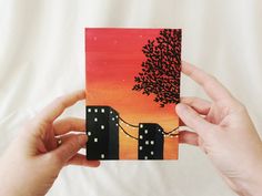 two hands holding up a piece of art that looks like cityscape at sunset