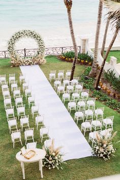 an outdoor ceremony setup with white chairs and flowers on the grass near the beach, surrounded by palm trees