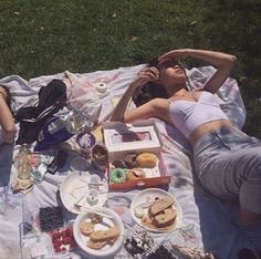 two women laying on a blanket in the grass with food and drinks laid out around them