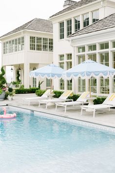 an empty swimming pool with lawn chairs and umbrellas near the side of a large white house