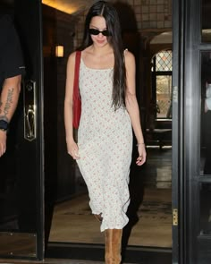 a woman in a white dress and brown boots is walking out of a building with her hand on her hip