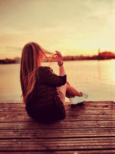 a woman sitting on a dock with her hair blowing in the wind and drinking water