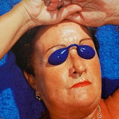 an older woman with blue eye patches on her eyes and hands over her head, in the sun