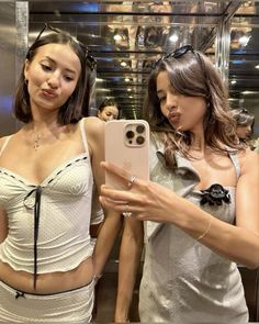 two beautiful women standing next to each other in front of a mirror holding an iphone