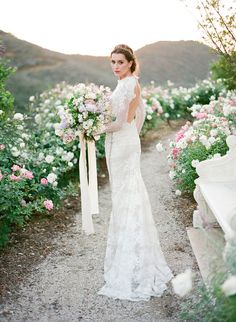 a woman in a wedding dress standing on a dirt road holding a bouquet of flowers