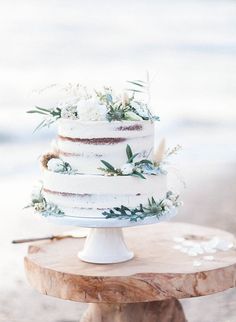 a three tiered cake with white flowers and greenery sits on a wooden stand