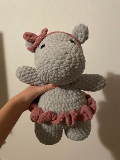 a hand holding a crocheted gray and pink hippo stuffed animal in front of a white wall