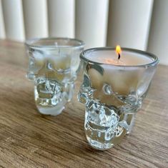 two glass candles sitting on top of a wooden table next to each other with a lit candle in the middle