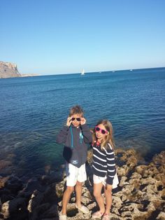 two young children standing on rocks near the ocean taking pictures with their cell phones while wearing sunglasses