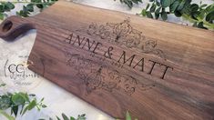 a wooden cutting board with the name and date engraved on it, surrounded by greenery