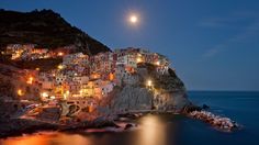 the moon is setting over an italian village by the water's edge, with its lights on