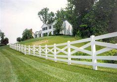 a white picket fence in front of a large house on a hill with trees and grass